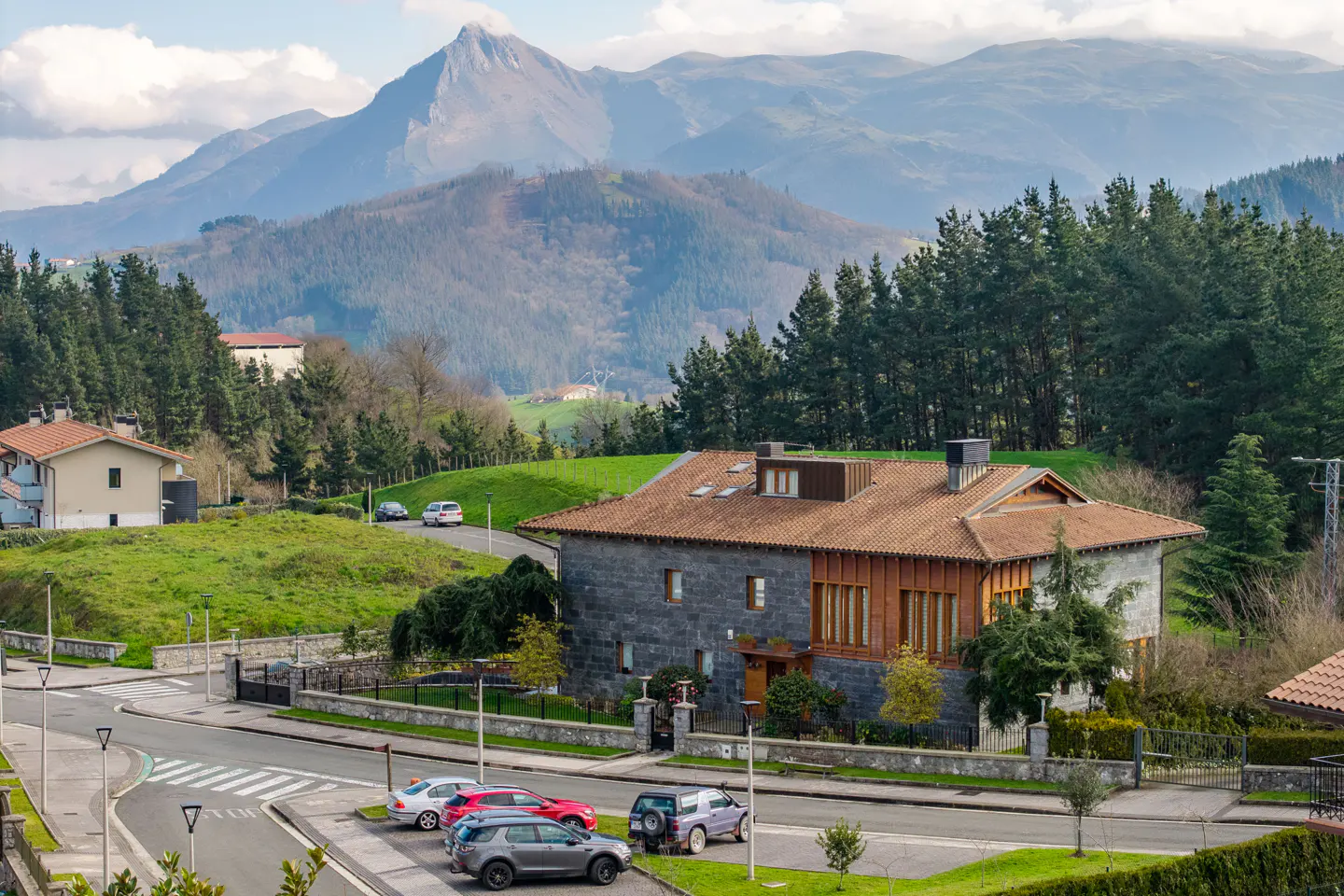A stone house with a red tile roof sits near a road with parked cars, with mountains in the background.