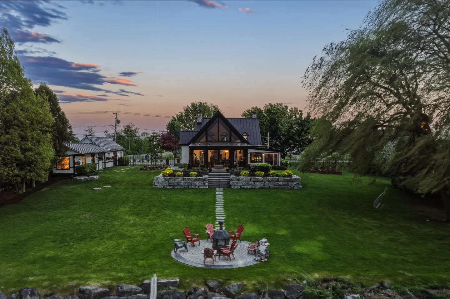 A modern farmhouse with a black roof and large windows sits on a green lawn with a stone path leading to a fire pit with chairs.