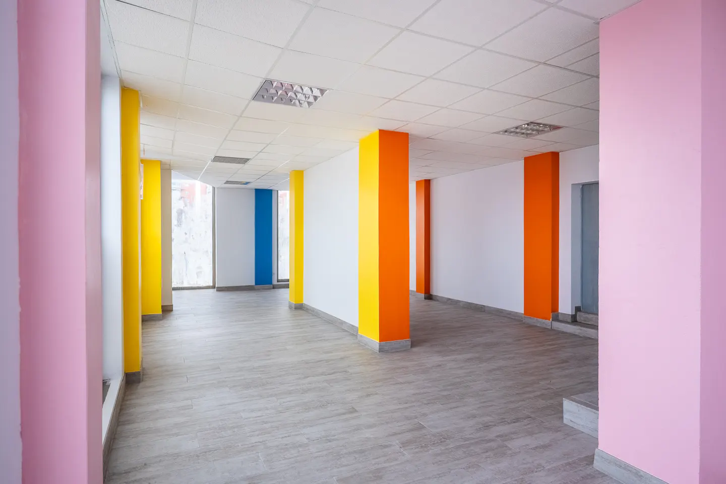 Empty office space with pink, yellow, orange, and blue columns. Gray wood-look floors and white walls.