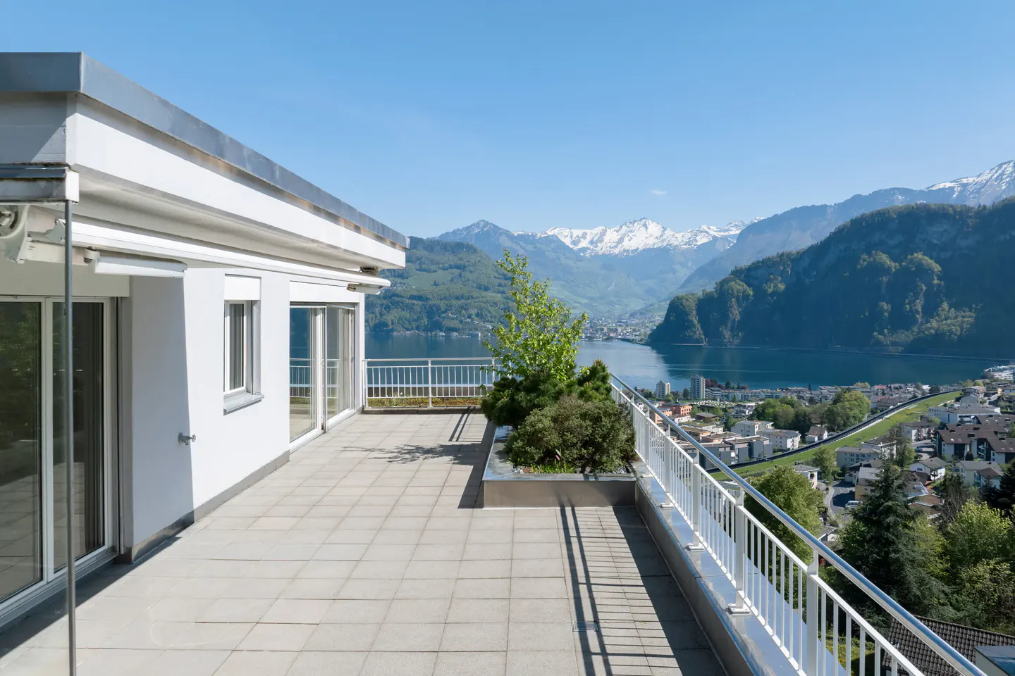A bright, sunny view from a modern apartment balcony with white railings, overlooking a lake, town, and snow-capped mountains.