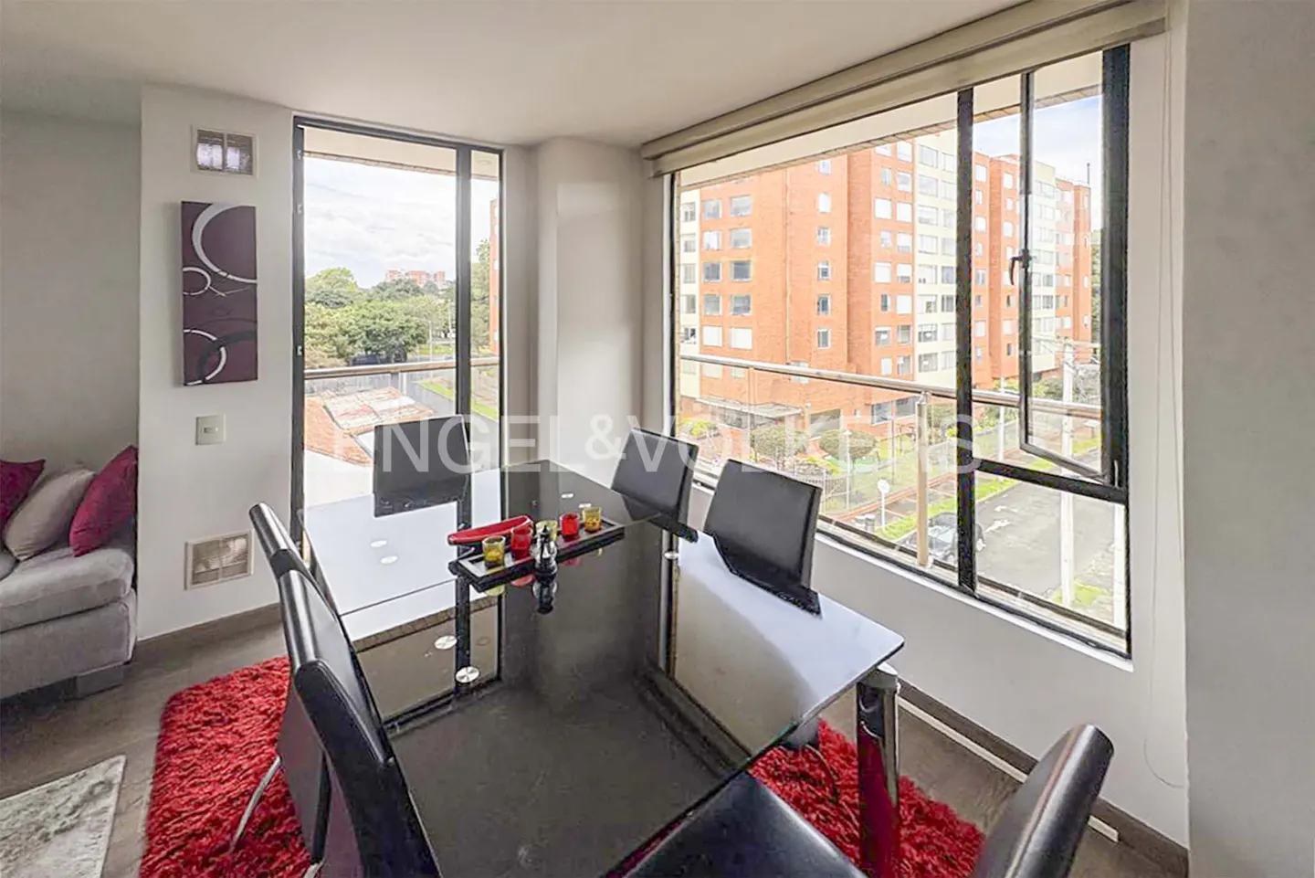 A modern dining room features a black glass table, black chairs, and a red rug. Large windows offer a city view.