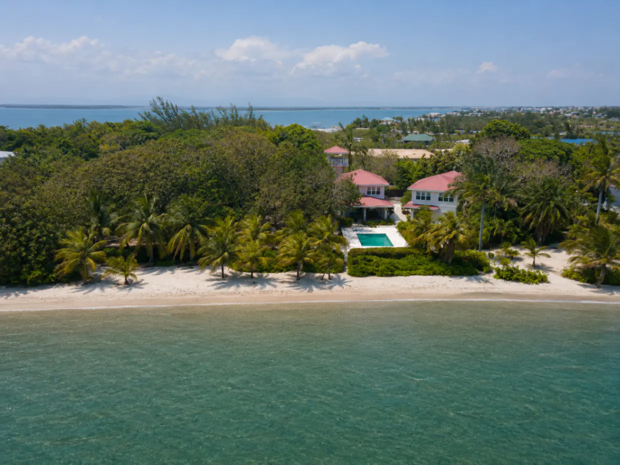 Aerial view of a beachfront property with white houses, pink roofs, a pool, and lush green trees. The turquoise ocean meets a sandy beach under a blue sky.