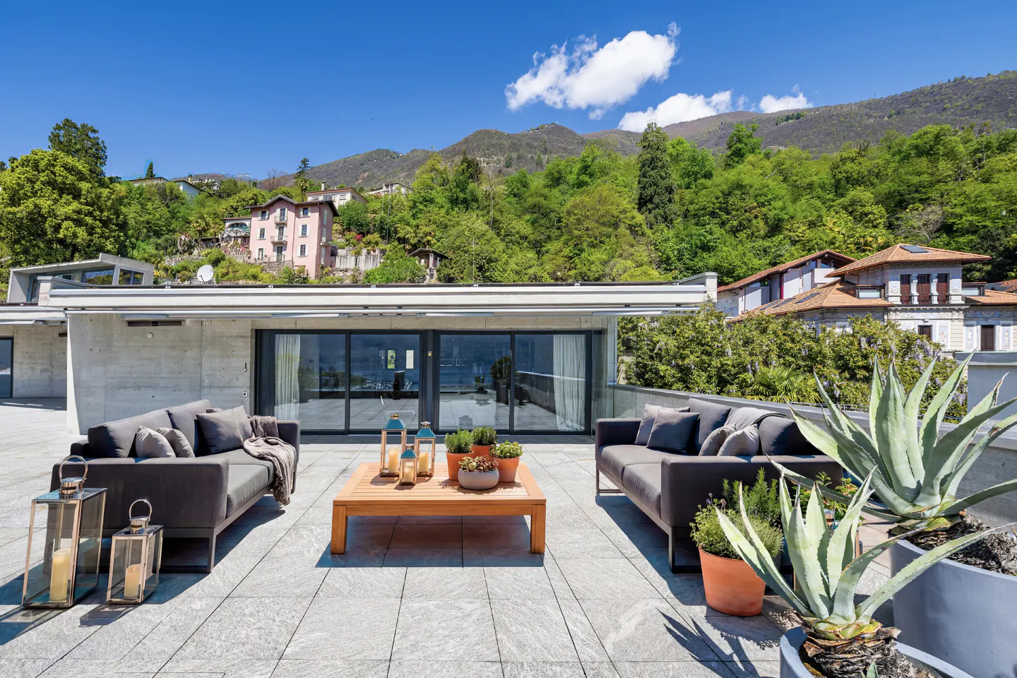 Luxury patio with gray sofas, wood table, and potted plants. Mountains and blue sky in the background.