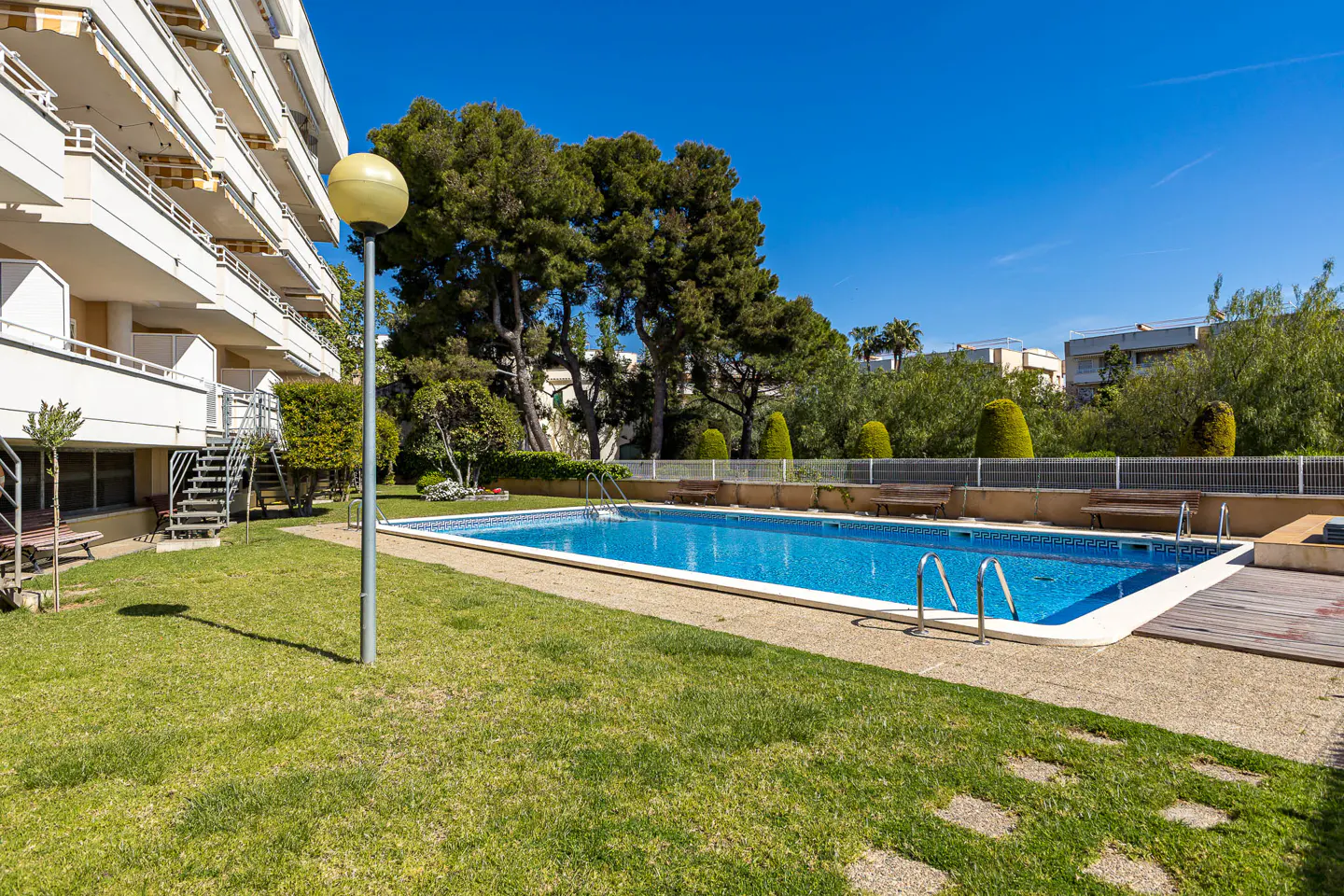 Outdoor pool with blue water, surrounded by green grass and trees, next to a white apartment building under a clear blue sky.