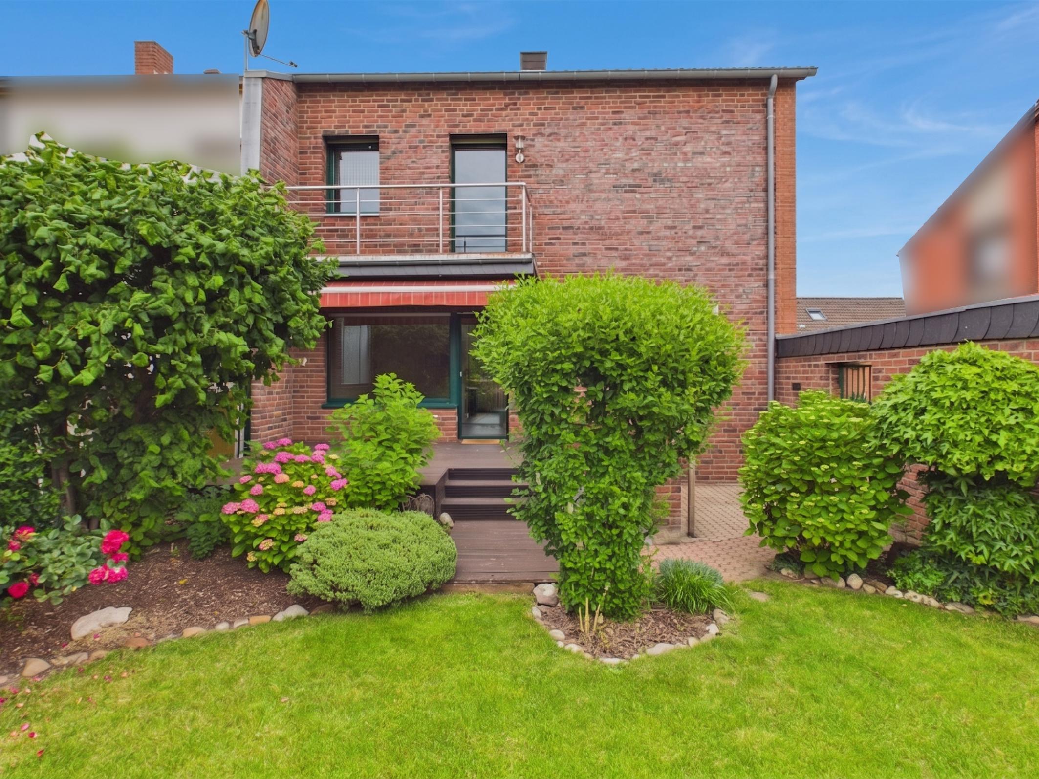 Red brick house exterior with a wooden deck, balcony, lush green bushes, pink flowers, and a manicured lawn under blue sky.