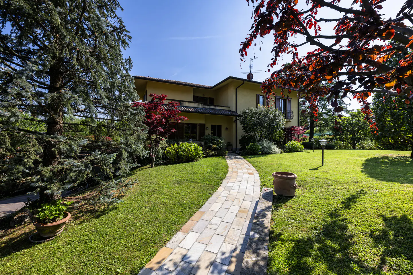 Two-story yellow house with a stone walkway leading through a green lawn, surrounded by trees and plants under a blue sky.