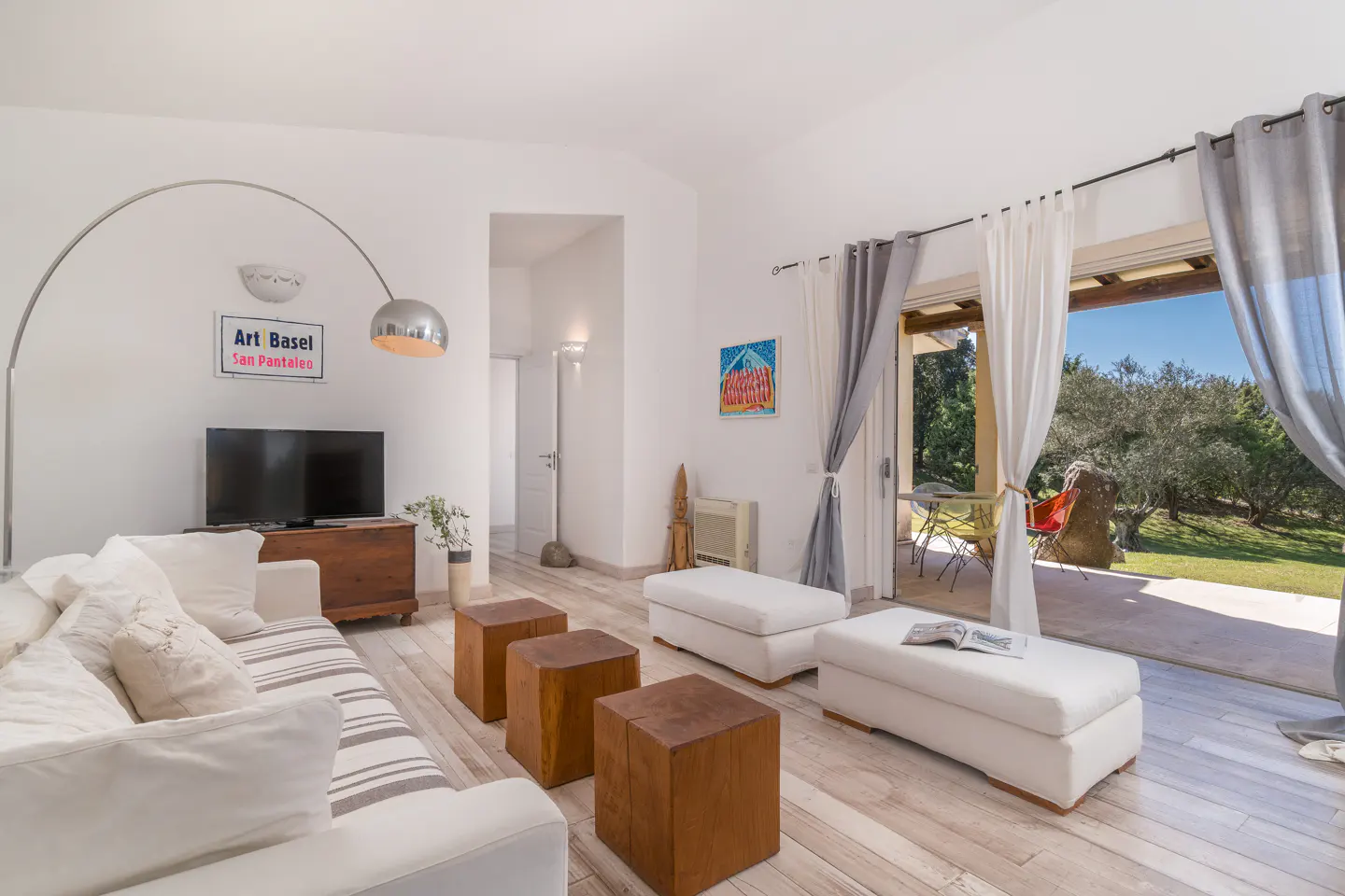Bright living room with white sofa, wooden cube tables, and patio doors to a green lawn. A silver arc lamp stands near a TV.