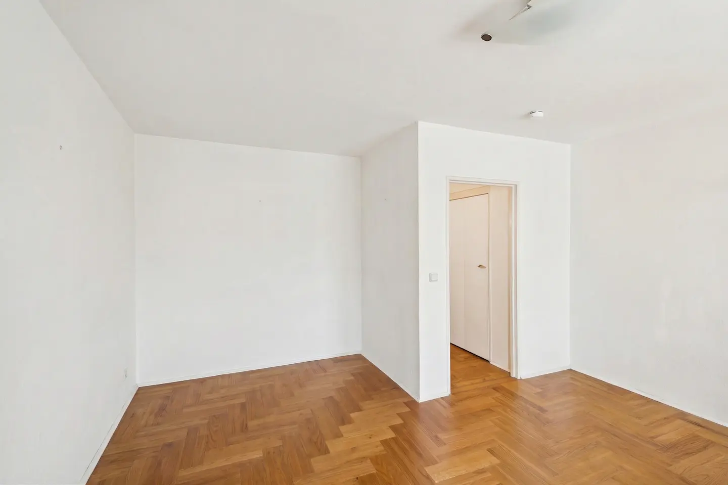 Empty room with white walls, herringbone wood floor, and an open doorway to another room.