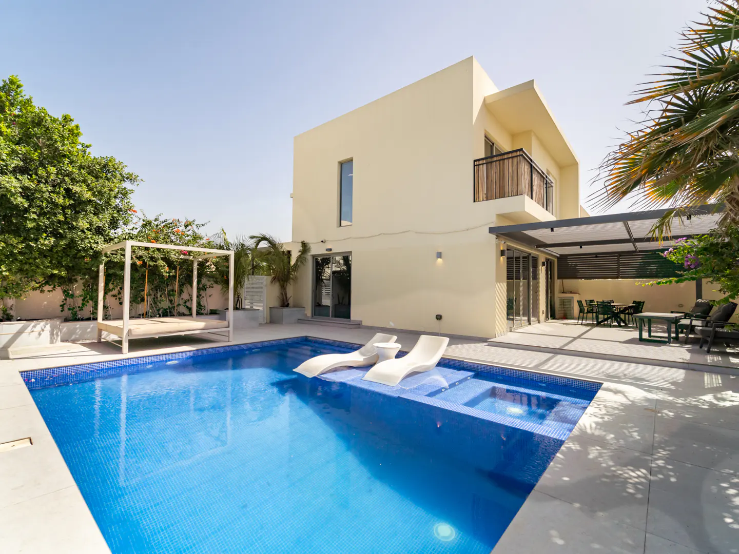 Exterior of a two-story beige house with a blue tiled pool, white lounge chairs, and a covered patio.