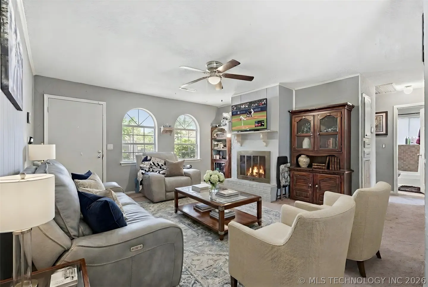 Living room with gray walls, a ceiling fan, and a fireplace with a TV above it. There is a gray sofa, two beige chairs, and a wooden coffee table.