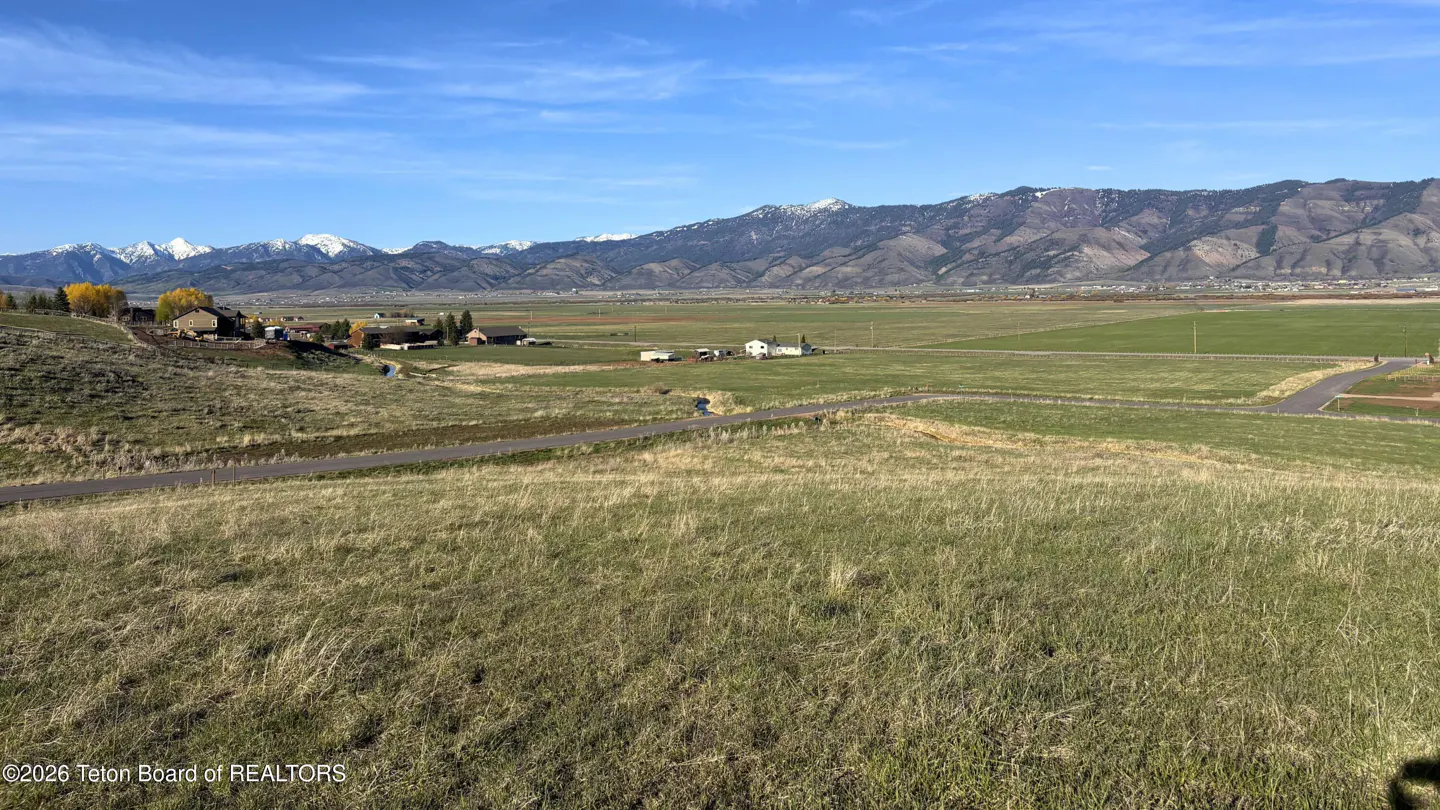 Scenic view of a grassy field with a road, houses, and snow-capped mountains against a blue sky.