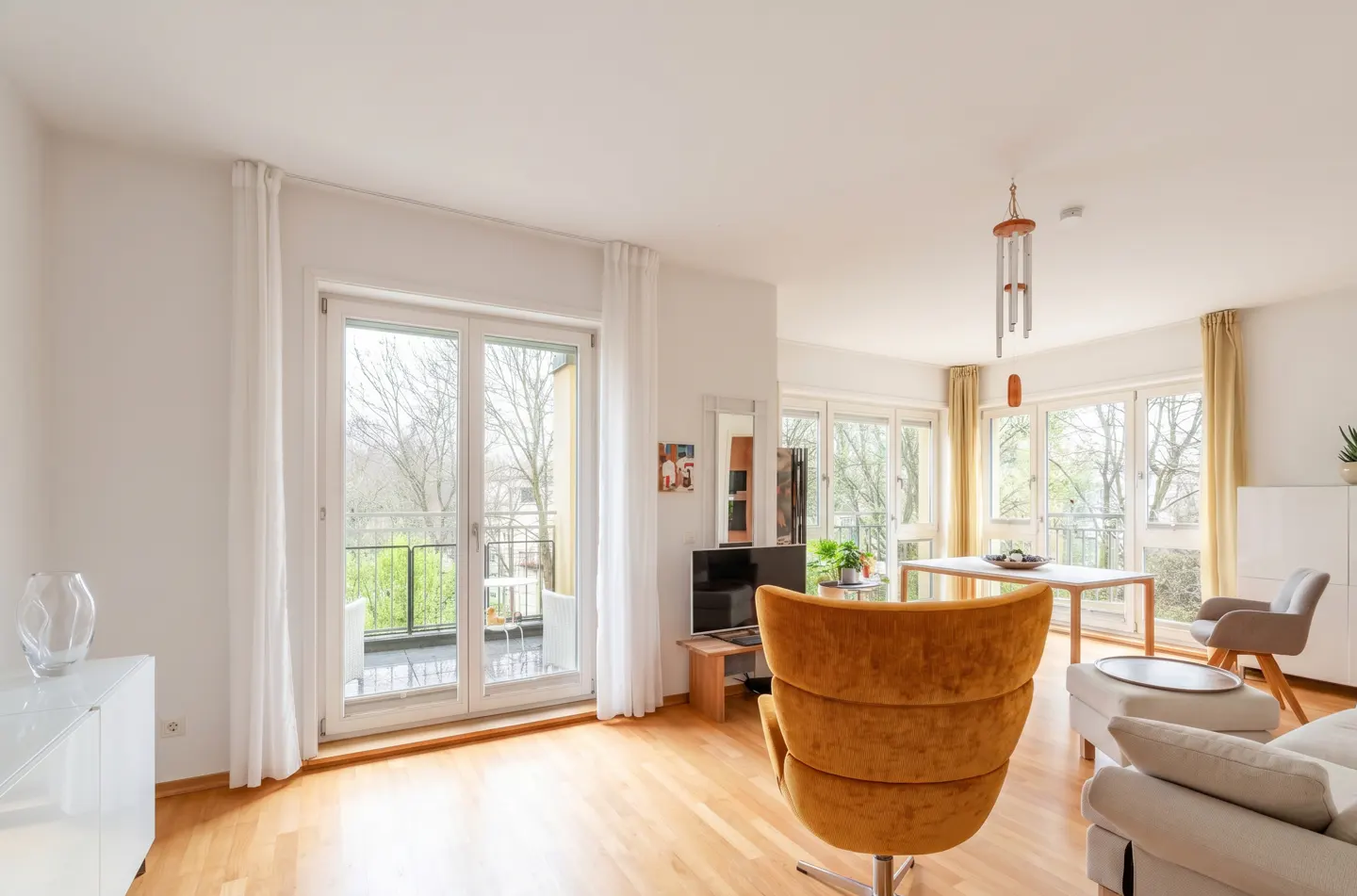 Bright living room with wood floors, white walls, and large windows. A yellow chair faces a balcony with a view of trees.