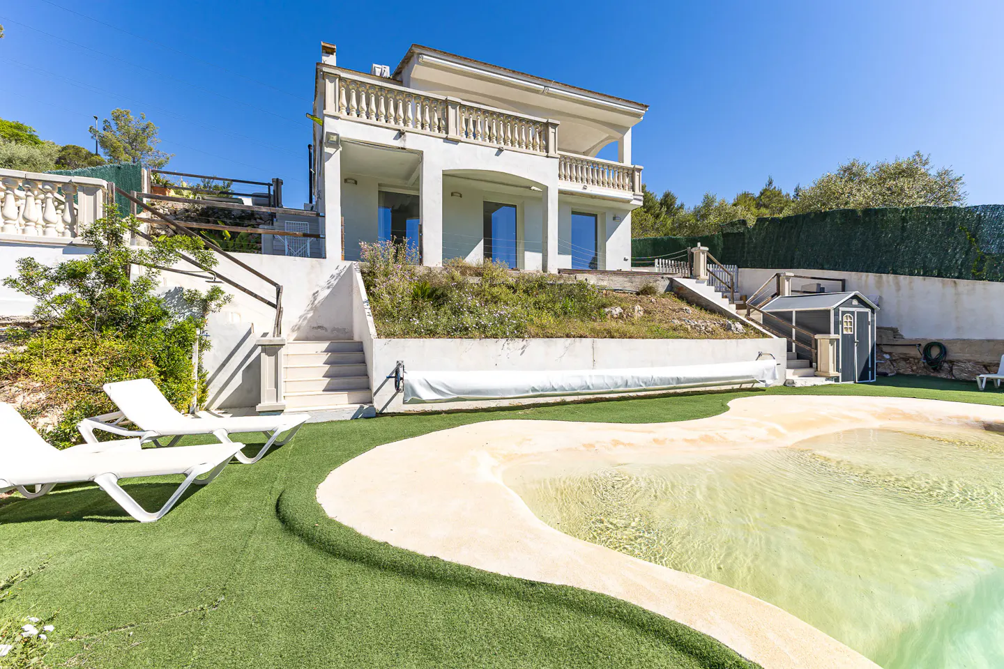 Two-story white house with a balcony overlooking a pool with a sandy beach area and green artificial turf. Two white lounge chairs are visible.