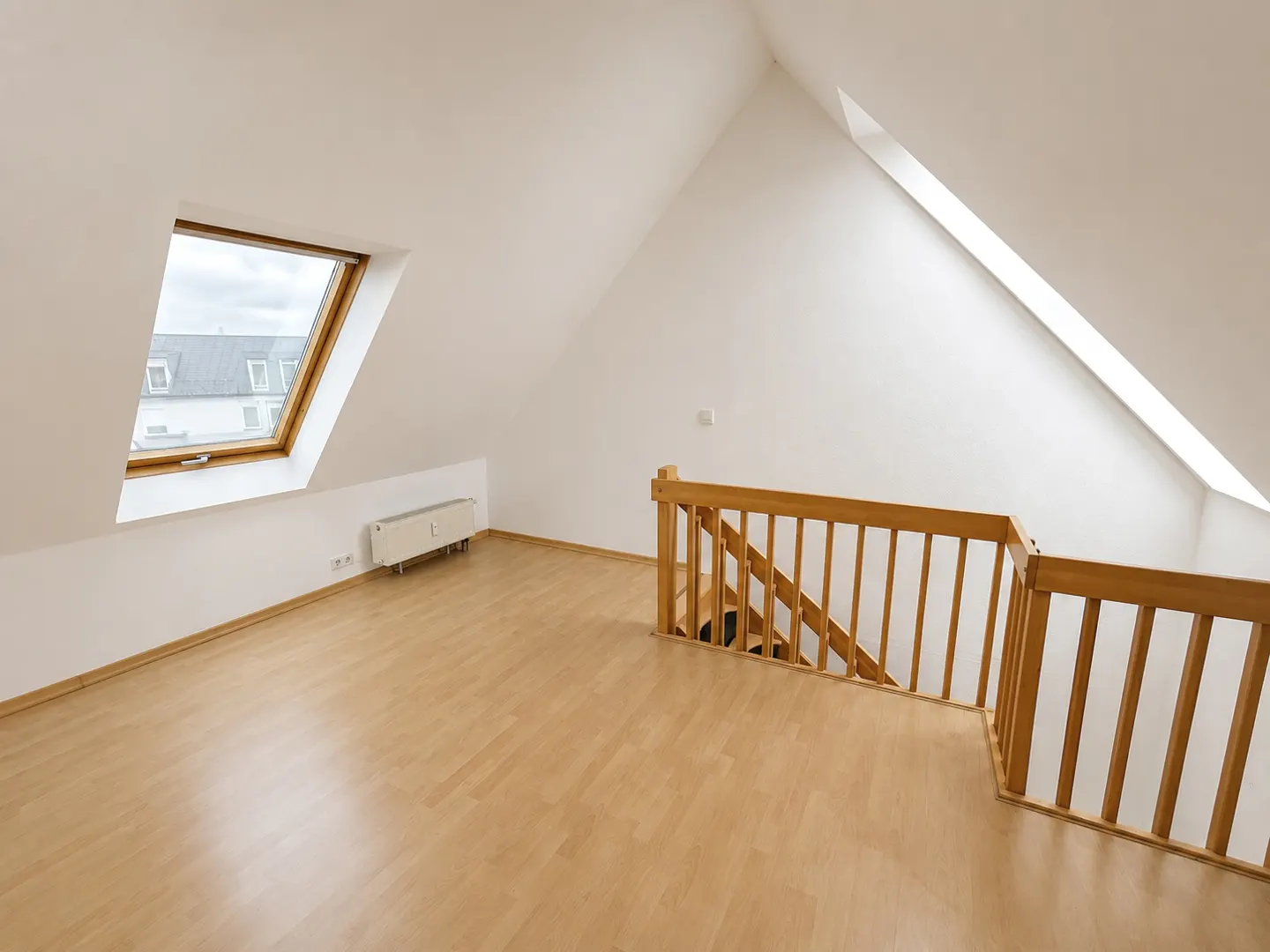 Attic room with light wood floors, white walls, and a wooden staircase railing. A window shows a view of rooftops.