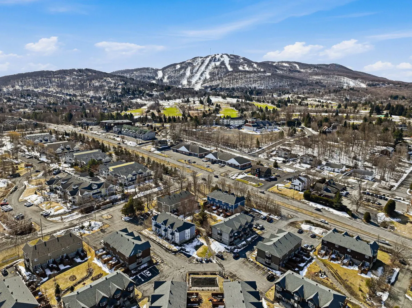 Aerial view of a residential area with houses, roads, and a snow-covered mountain in the background under a blue sky.