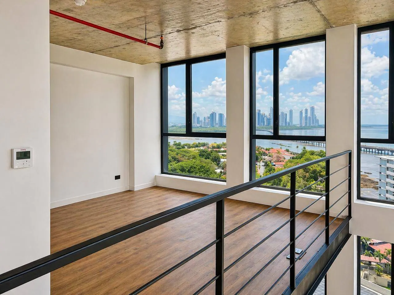 Modern loft interior with wood floors, white walls, and black framed windows. City skyline view with water and greenery visible. Black metal railing in foreground.