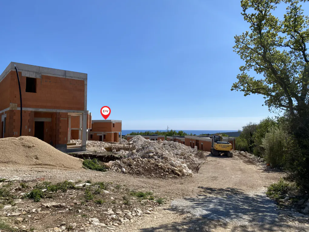 Construction site with unfinished brick buildings, piles of gravel, and a yellow excavator under a clear blue sky.