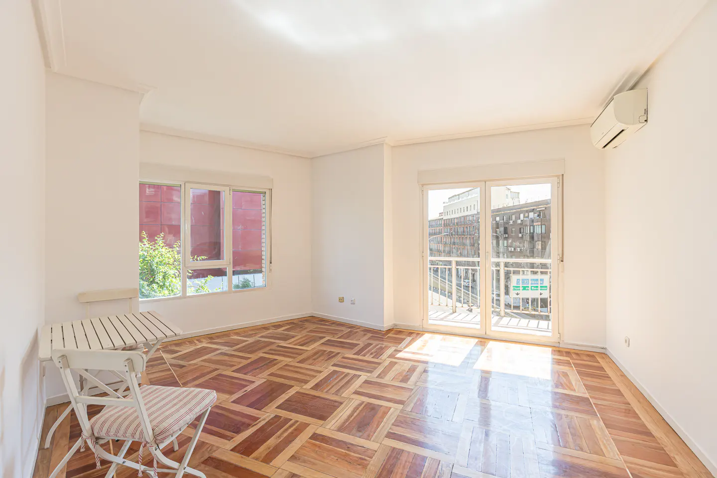 Bright, empty room with parquet floors, white walls, and a window and balcony door showing city views. A small white table and chair sit in the corner.