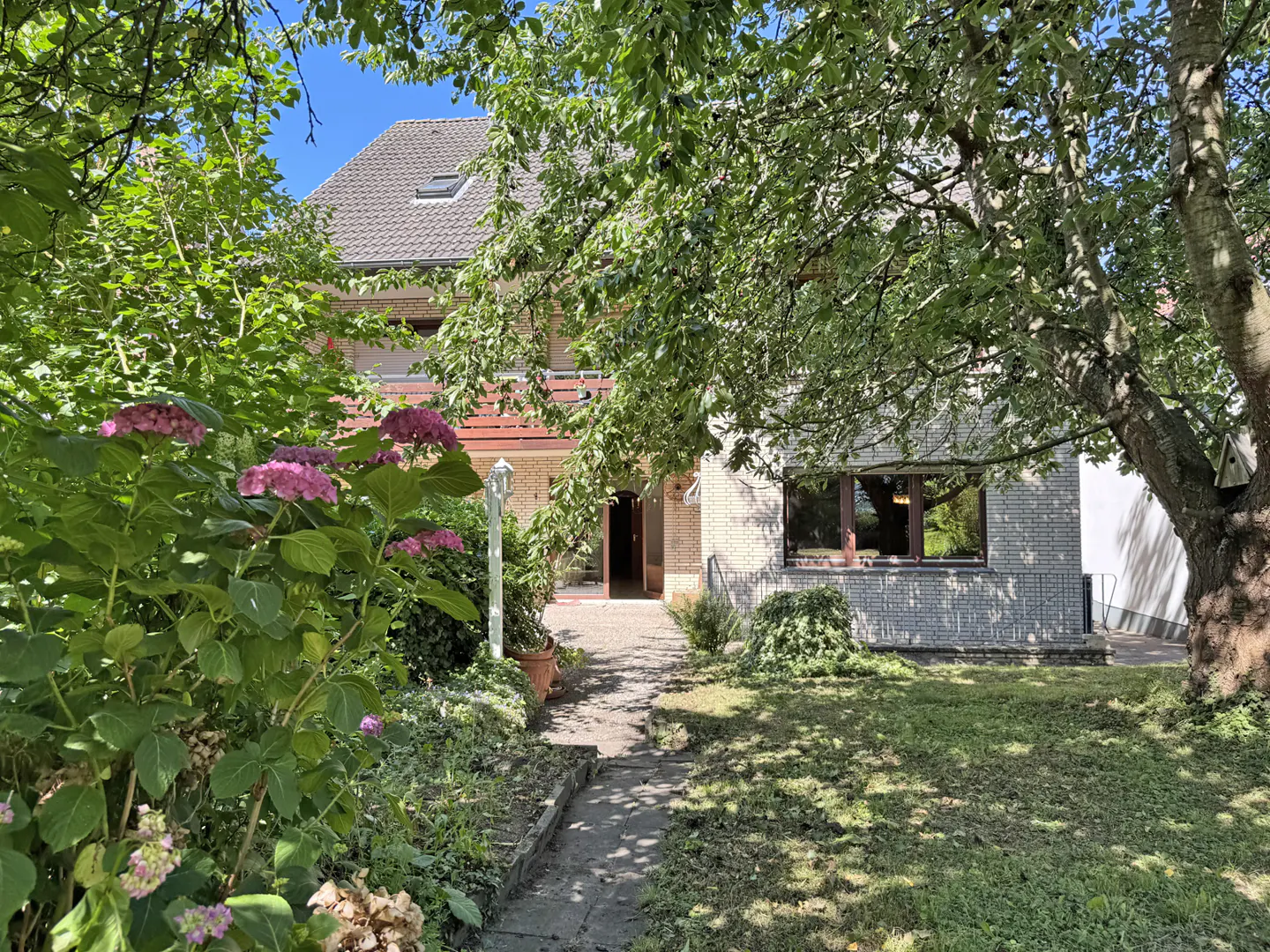 A light brick house with a balcony, viewed from a garden path framed by lush green trees and pink hydrangea bushes.