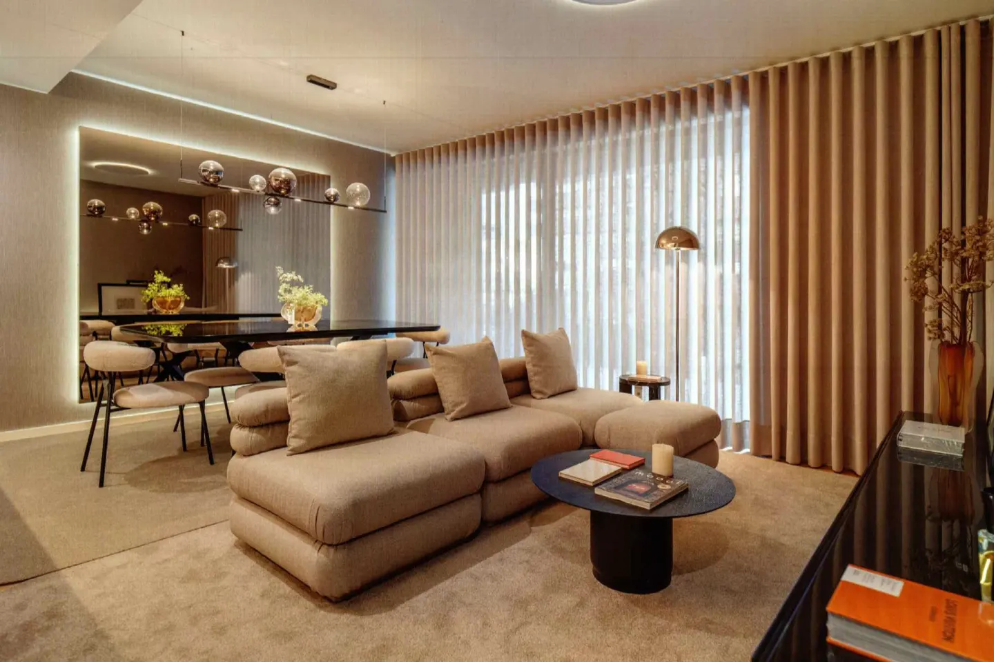 Living room with a beige sectional sofa, round black coffee table, and a dining table reflected in a large mirror. Curtains and a floor lamp are visible.