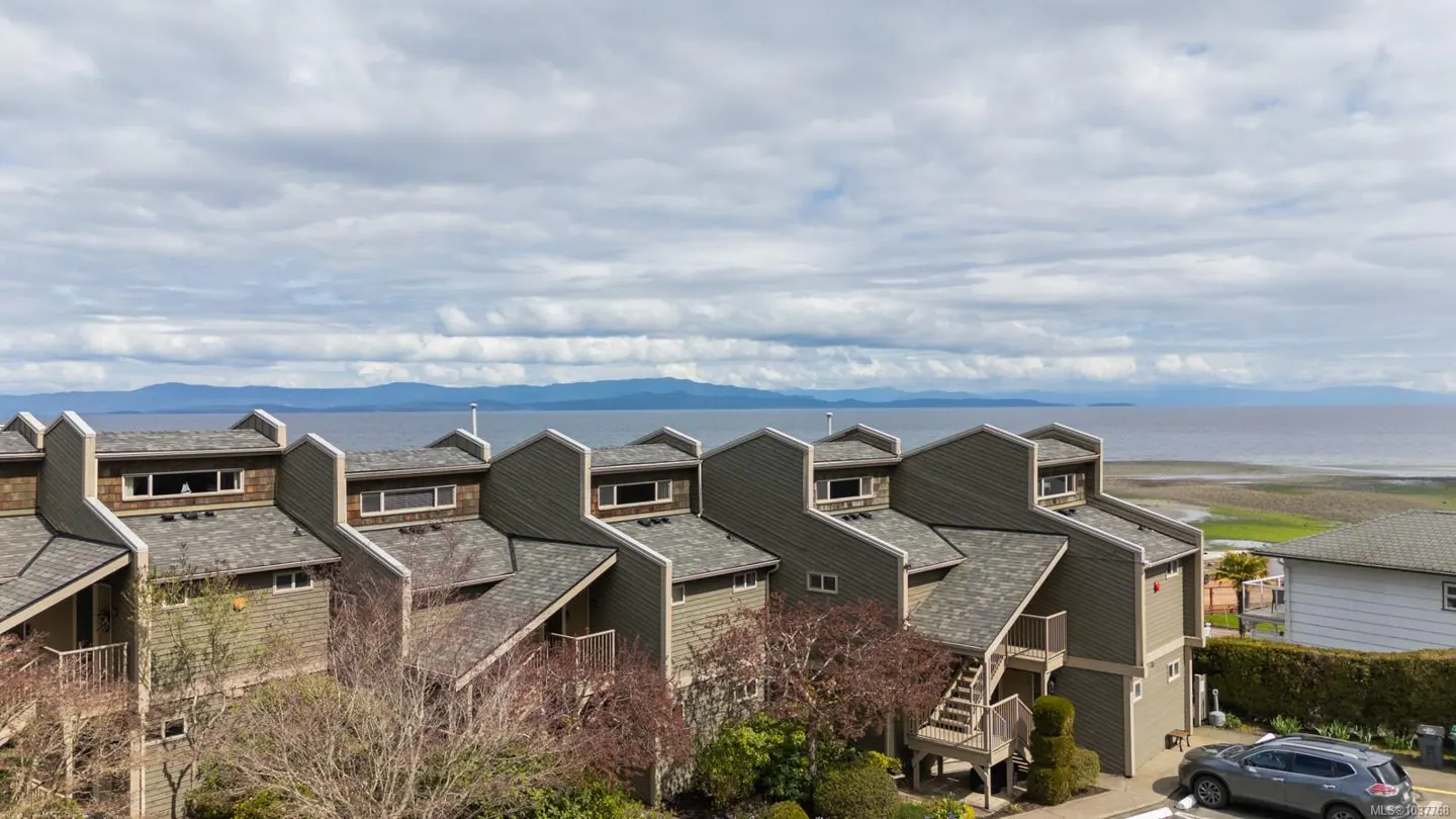 View of townhouses with gray roofs, green siding, and ocean view under a cloudy sky. A gray car is parked in front.