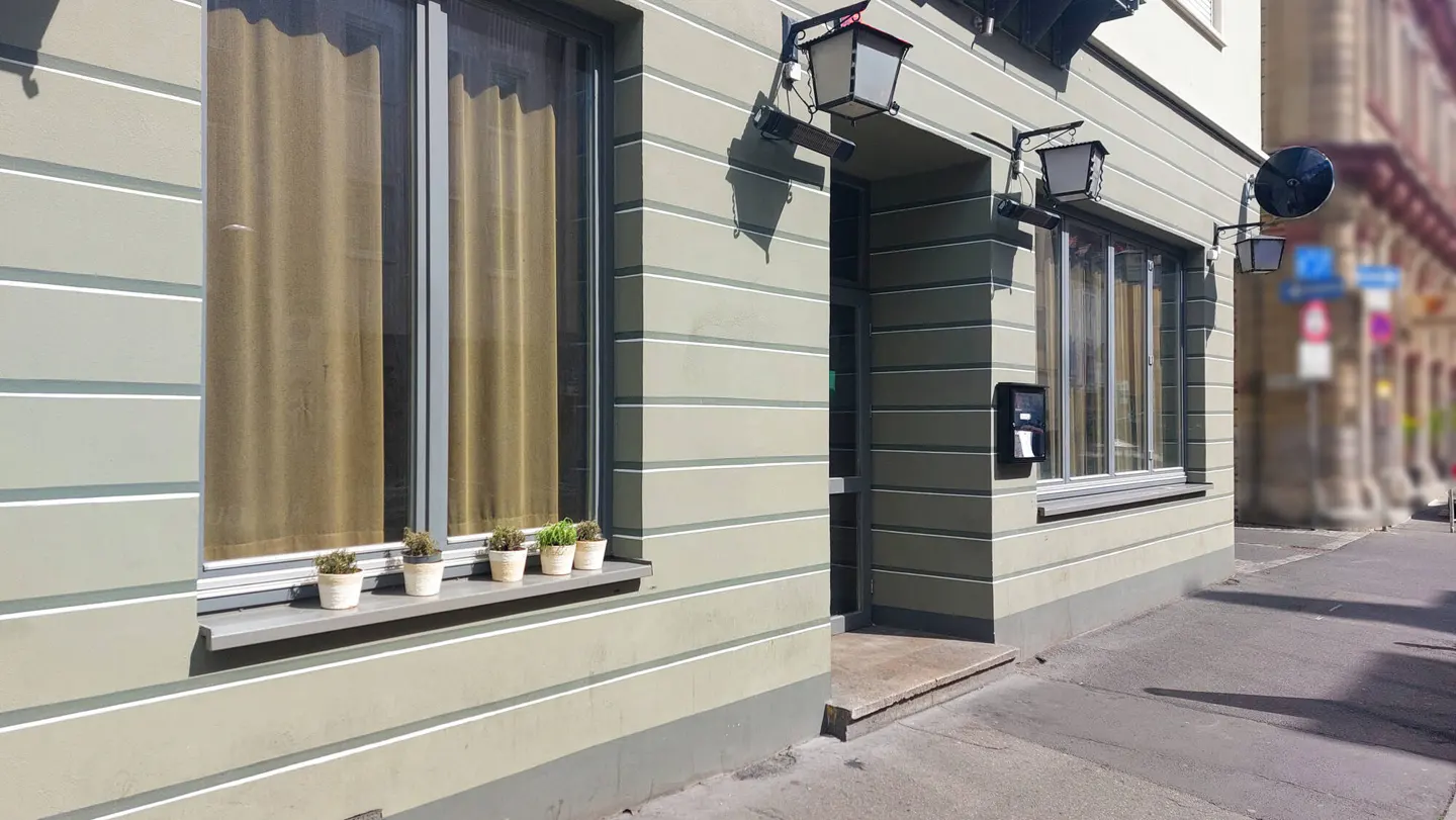 Exterior of a building with gray walls, white stripes, windows with curtains, and potted plants on the windowsill.