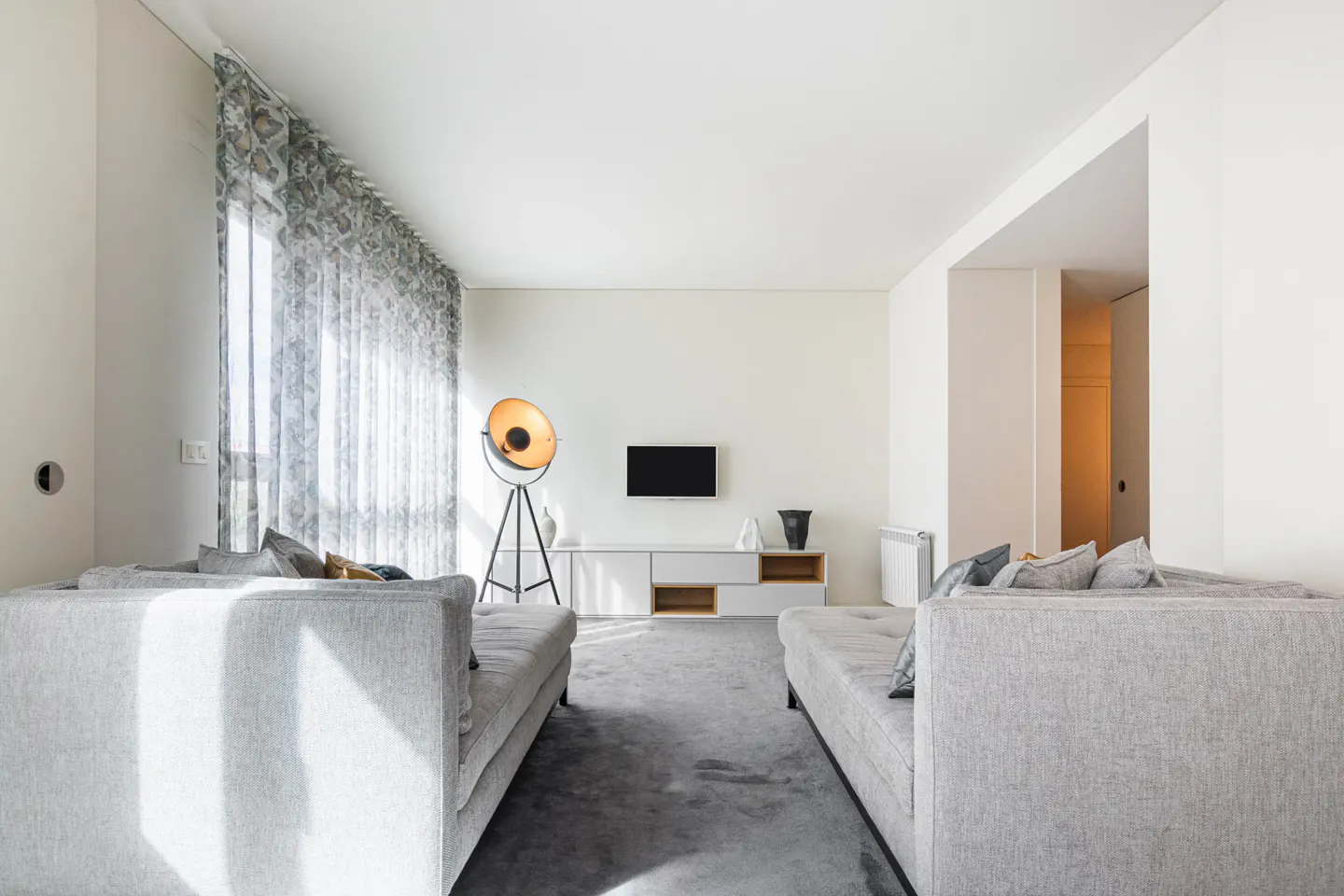 Bright living room with two gray sofas facing each other, a black TV, and a gold floor lamp. Gray carpet and white walls.