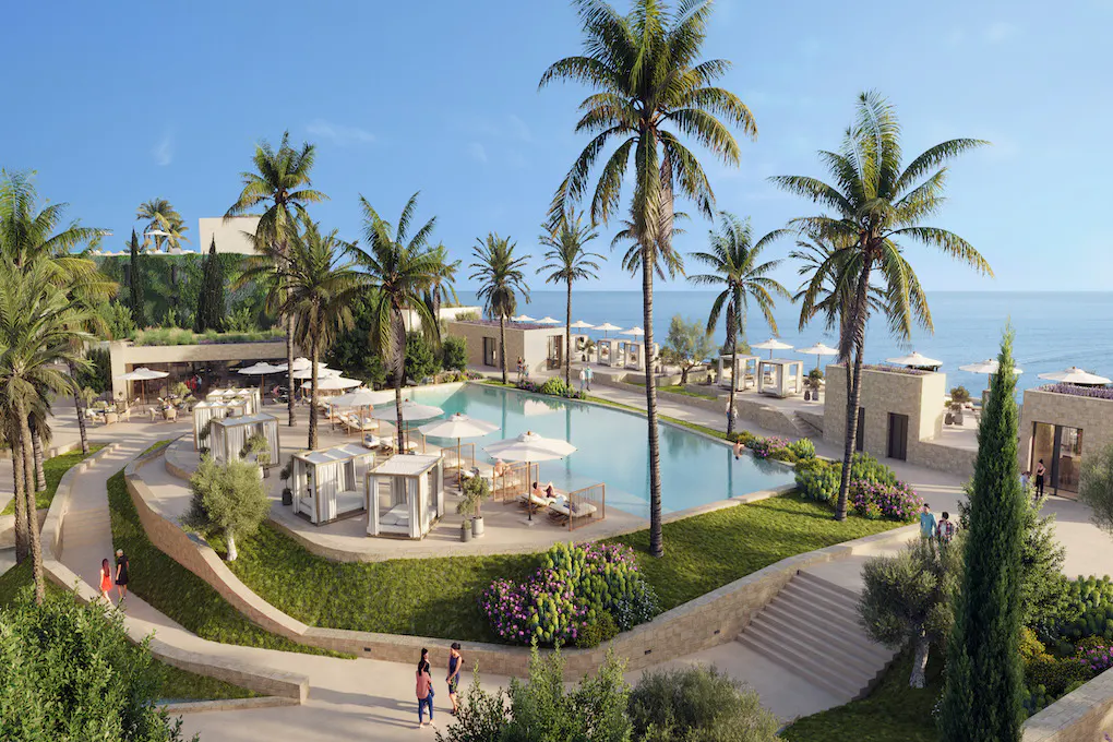 Aerial view of a luxury resort with a pool, palm trees, cabanas, and ocean views under a clear blue sky.