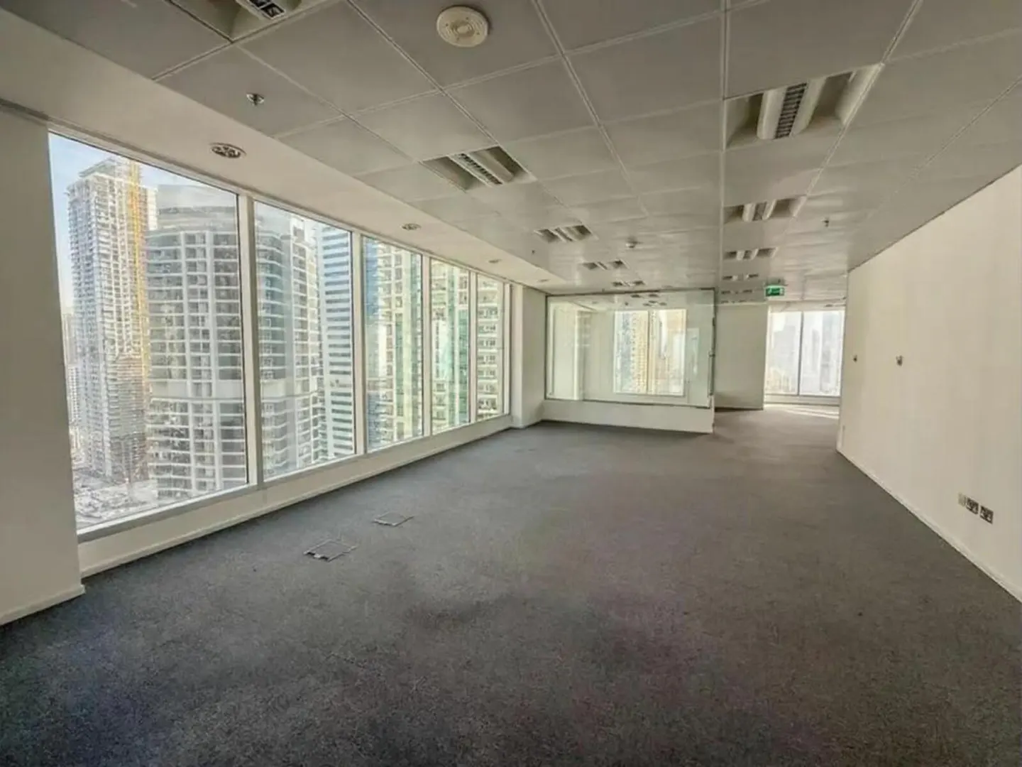 Empty office space with gray carpet, white walls, and a ceiling with recessed lighting. Large windows offer a city view.