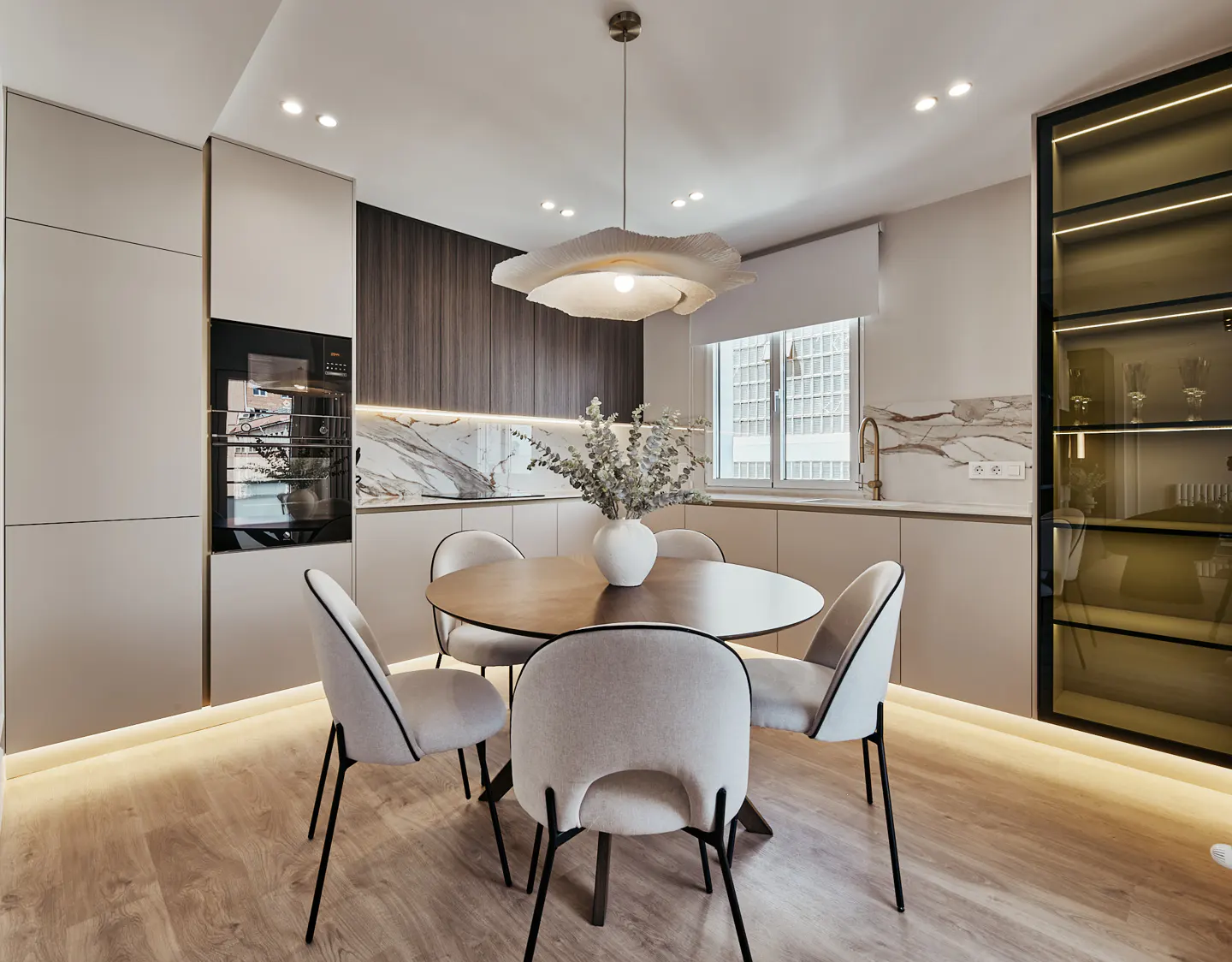 A modern kitchen with a round wooden table surrounded by four white chairs. A white vase with flowers sits on the table. A unique light fixture hangs above.