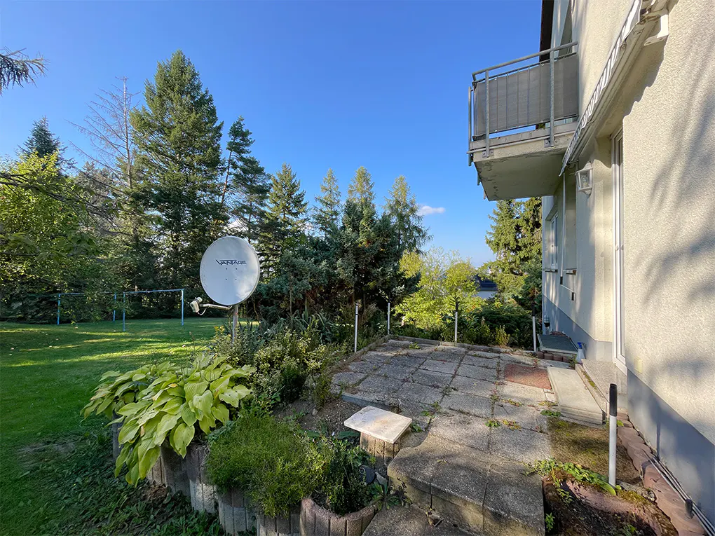 Exterior view of a home with a stone patio, lush green lawn, trees, and a satellite dish under a blue sky. A balcony is visible on the side of the house.