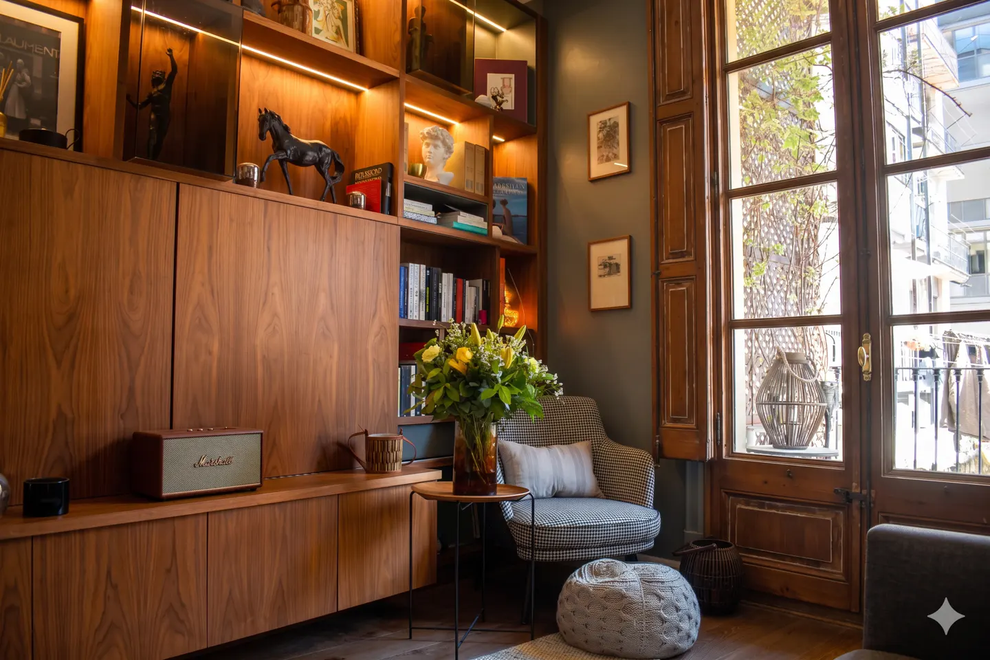 A cozy living room features a wooden cabinet with shelves, a houndstooth chair, and a large window. A vase of yellow flowers sits on a small table.