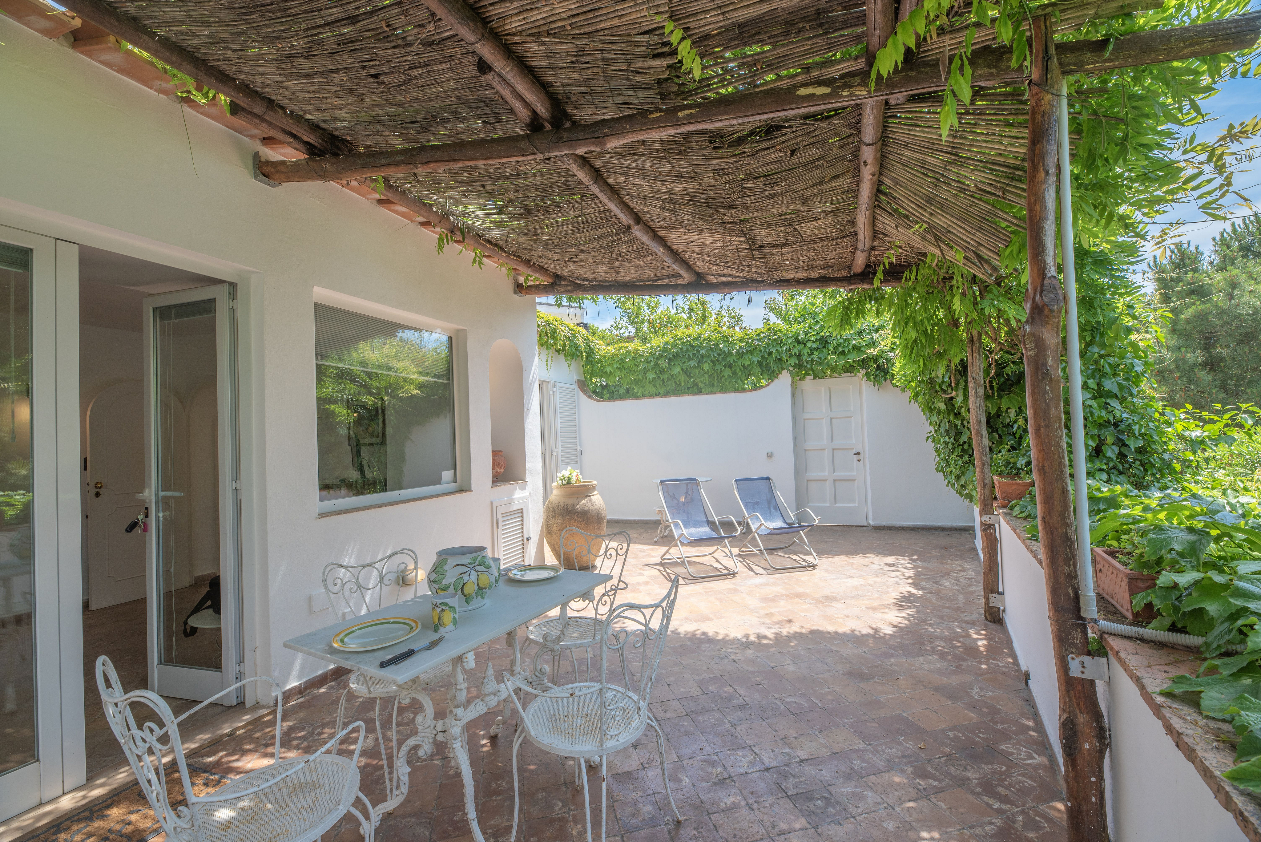 Outdoor patio with white table and chairs set for dining, under a thatched roof. Lush greenery surrounds the space.