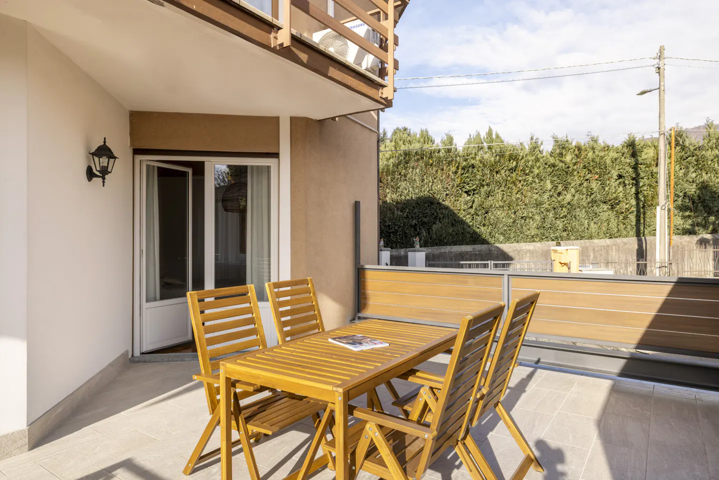 Outdoor patio with a wooden table and chairs, a magazine on the table, and a view of a green hedge in the background.