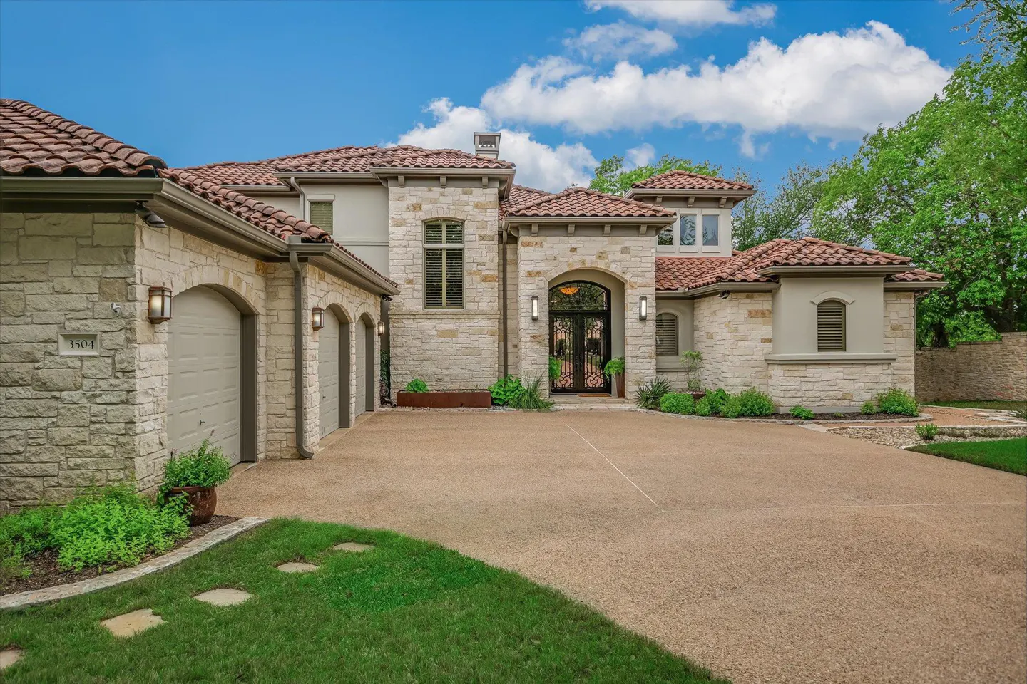 A tan stone house with a red tile roof, black double doors, and a three-car garage.