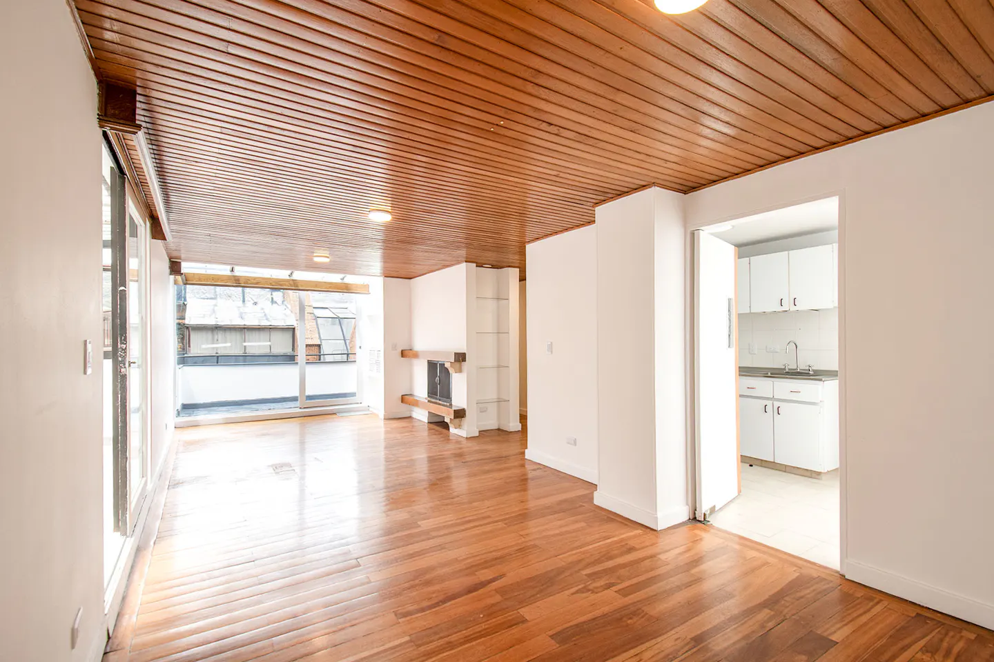 Bright living room with wood-paneled ceiling, hardwood floors, fireplace, and a view of a balcony, with a kitchen adjacent.