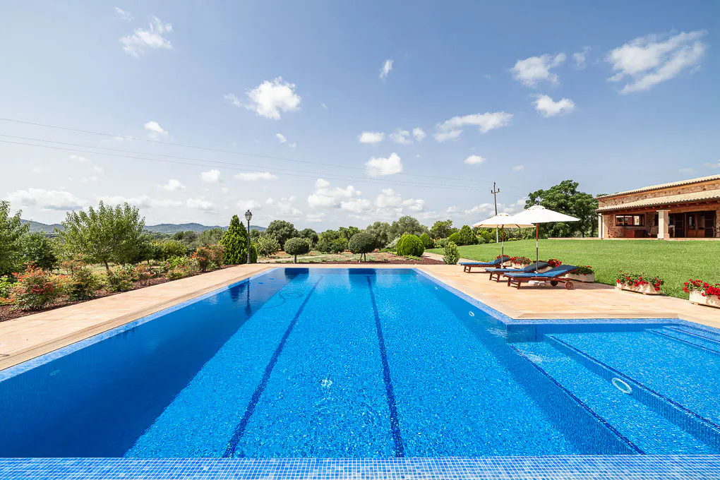 A bright blue tiled pool with lounge chairs and a white umbrella sits on a green lawn near a stone house under a blue sky.