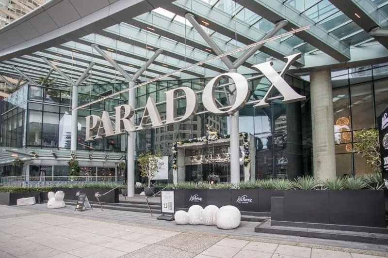 Exterior shot of the Paradox Hotel with a glass canopy and large white "PARADOX" sign. Planters and modern seating are in front.