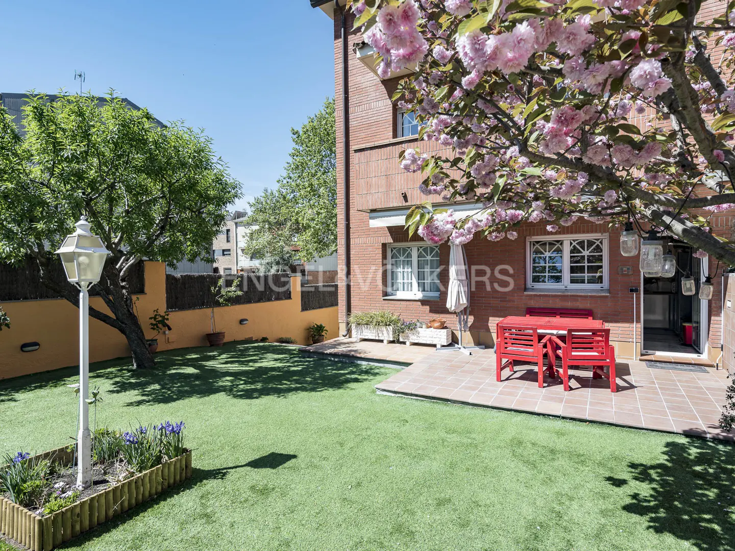 A brick house with a green lawn, a red table and chairs, and a flowering tree.