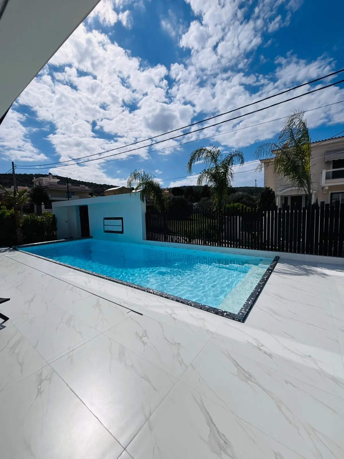 A bright blue swimming pool is surrounded by white marble tiles, with a black fence and palm trees in the background.
