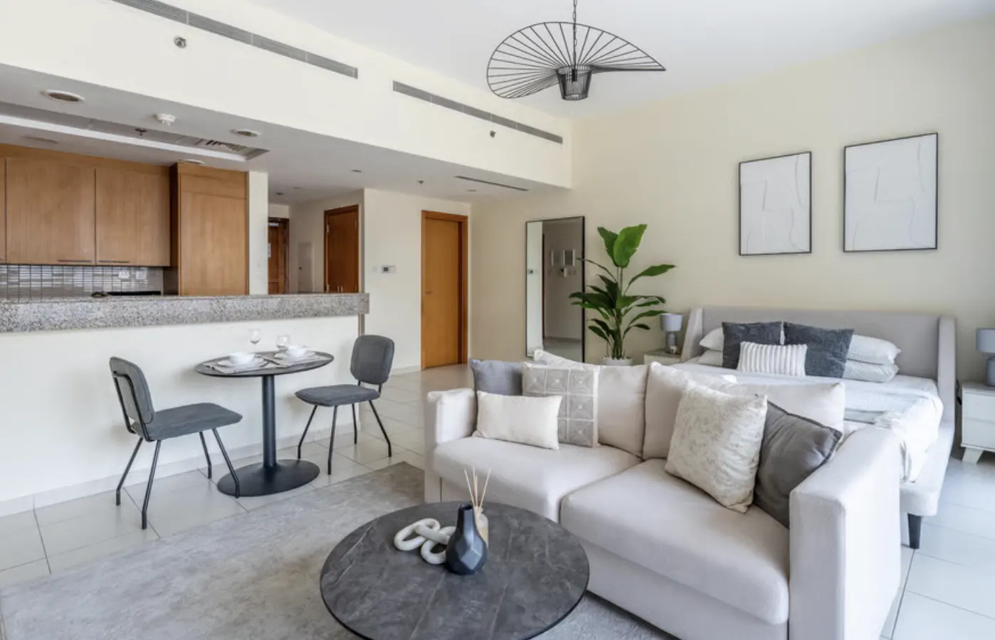 Studio apartment with a kitchen, dining table, sofa, and bed. The color scheme is neutral with white walls and light-colored furniture.