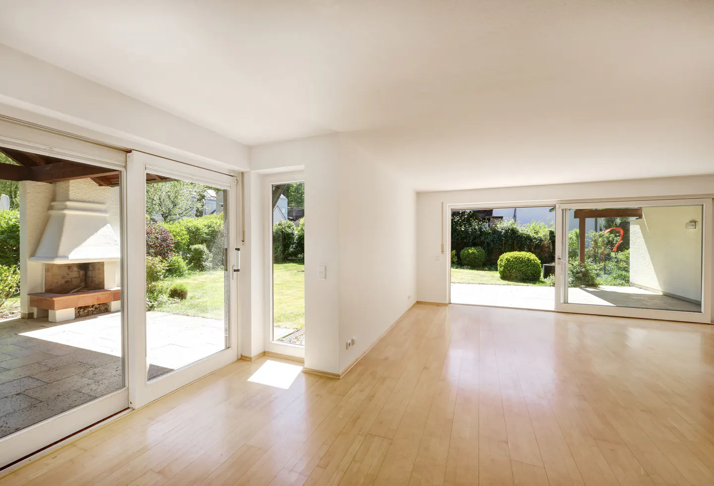 Bright, empty room with light wood floors and white walls. Sliding glass doors open to a patio with a fireplace and green yard.