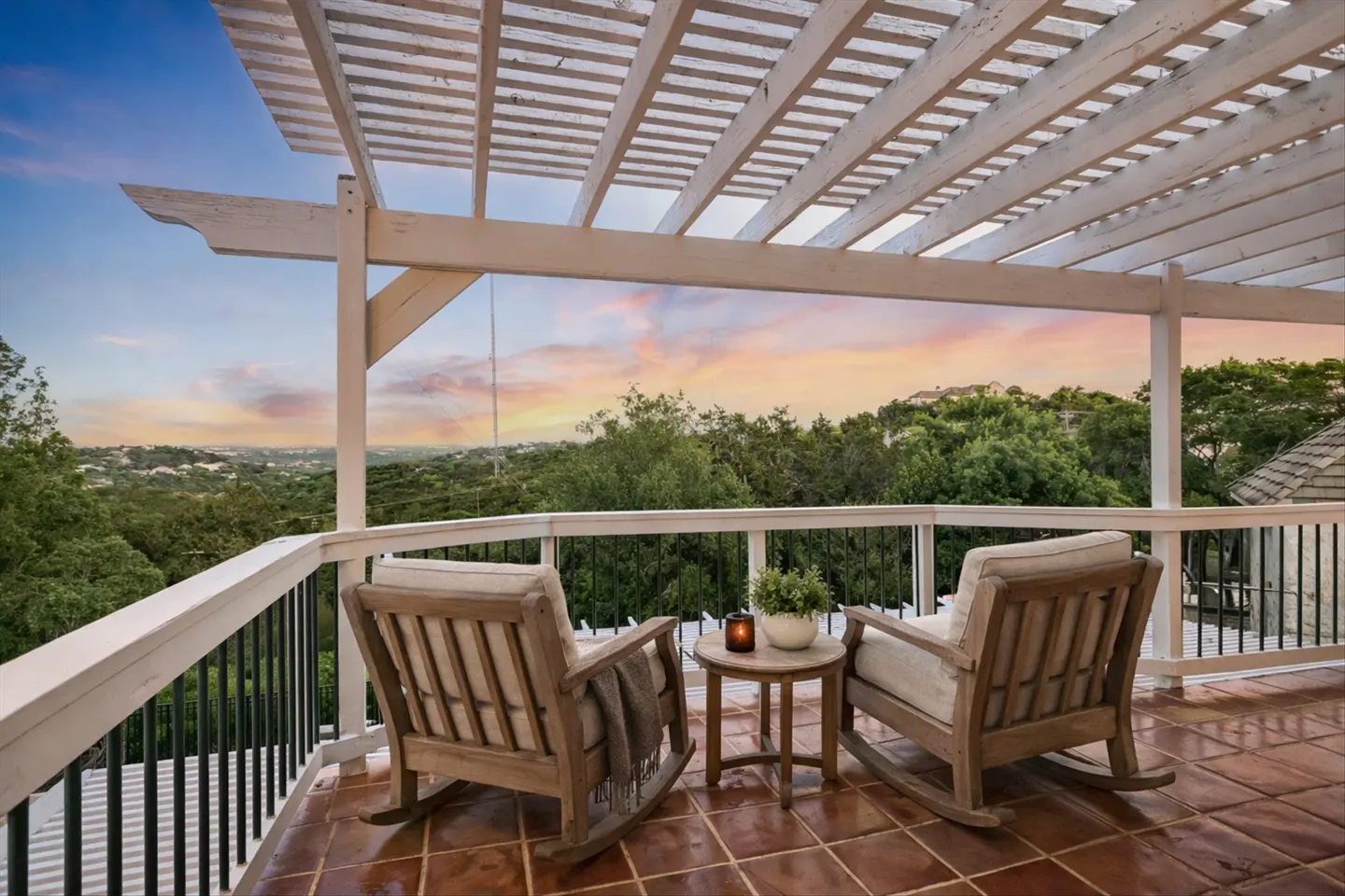 Outdoor patio with two rocking chairs, a small table, and a view of trees and a colorful sunset.