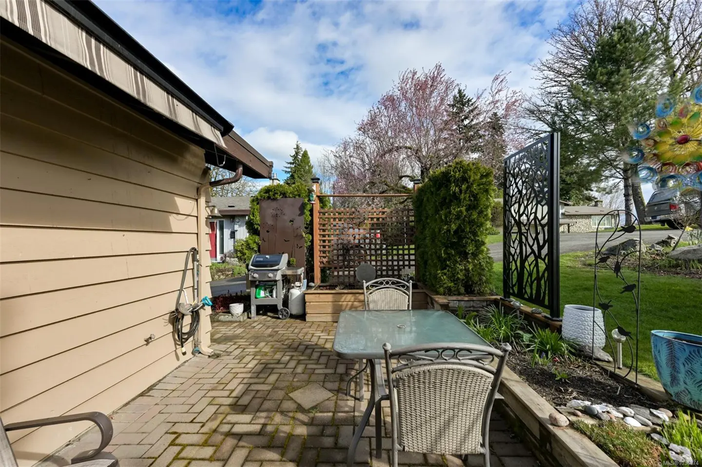 Outdoor patio with brick pavers, table, chairs, grill, and garden. The house has tan siding and a black metal screen.
