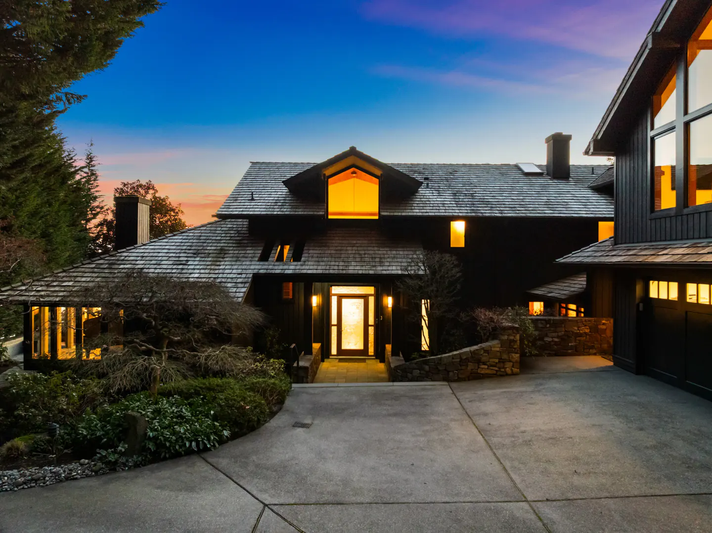 Exterior of a dark, two-story house with a gray shingle roof and a concrete driveway at dusk.