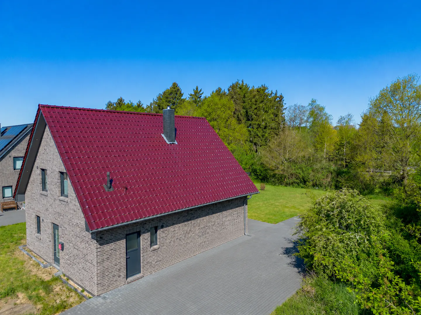 Aerial view of a brick house with a red tile roof, a gray chimney, and a paved driveway, set against a backdrop of green trees and a clear blue sky.