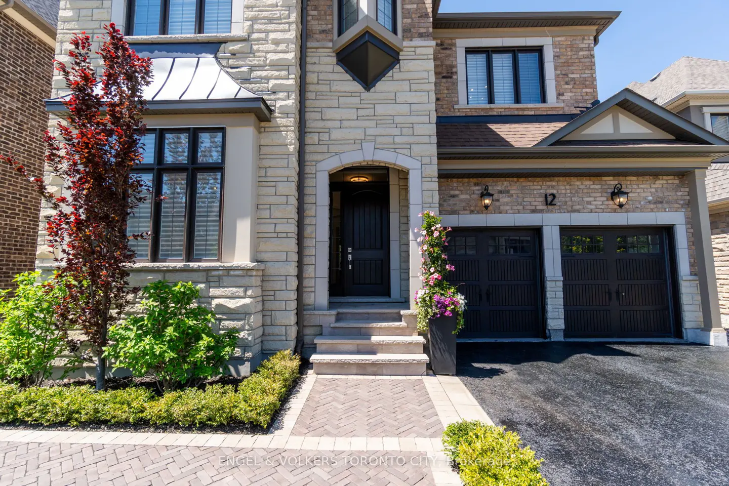 Two-story stone house with a dark front door, two-car garage, and a herringbone brick walkway under a clear blue sky.