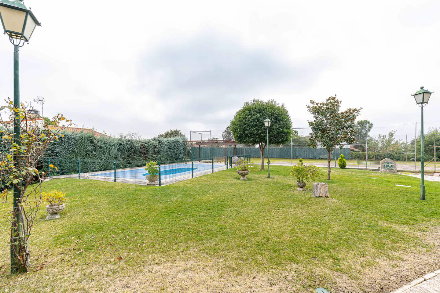 A backyard with a pool, green grass, trees, and lampposts under a cloudy sky.