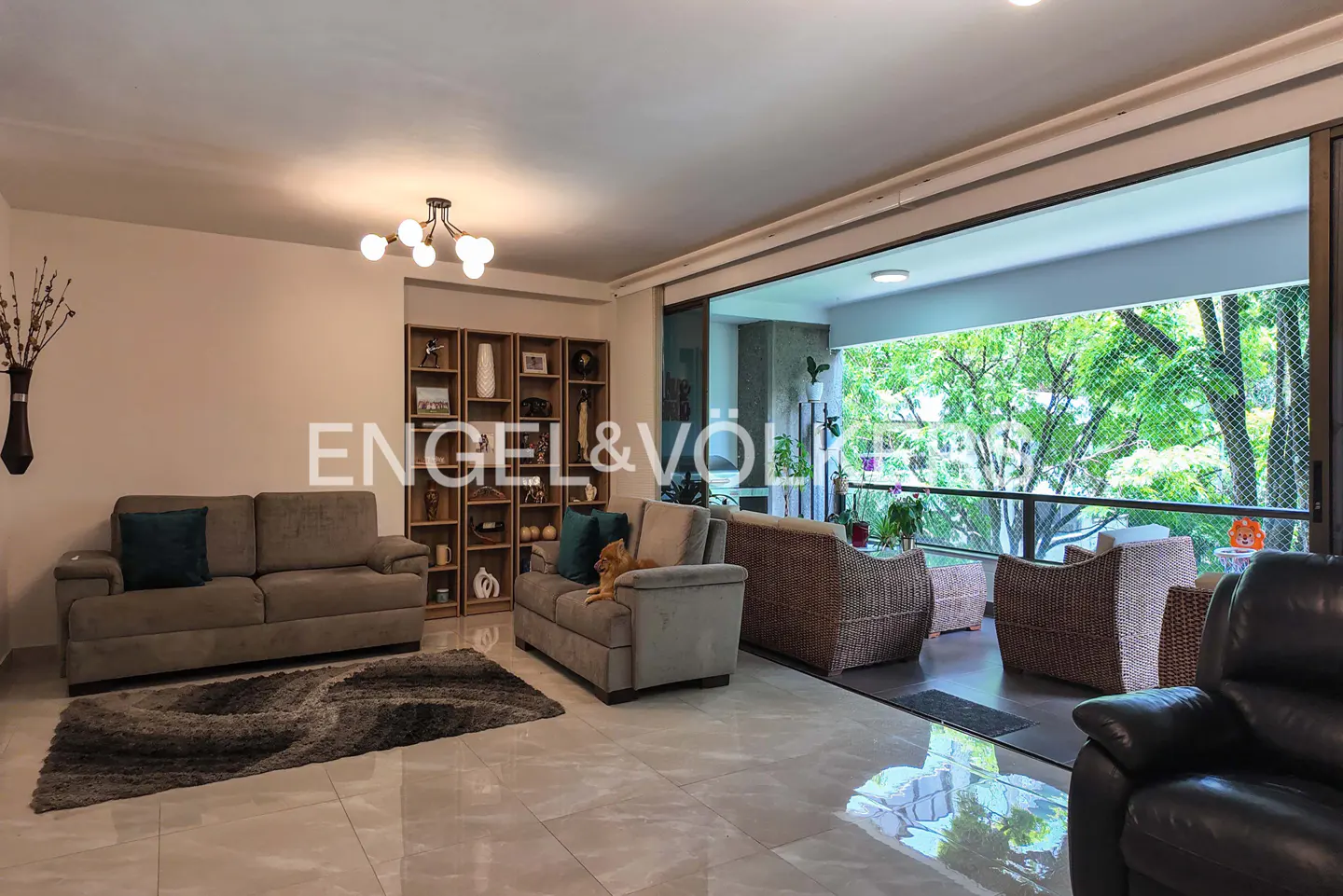Bright living room with sofas, rug, and open balcony with wicker furniture overlooking lush greenery.