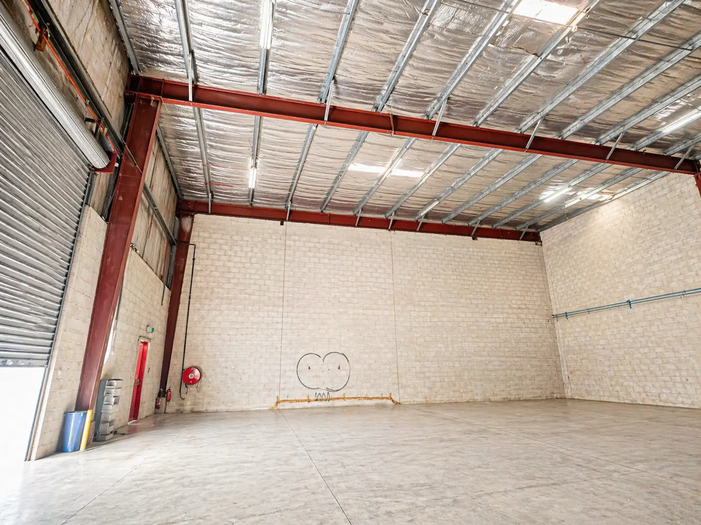 Empty warehouse interior with concrete floor, brick walls, and metal ceiling supports. A red fire extinguisher is mounted on the wall.